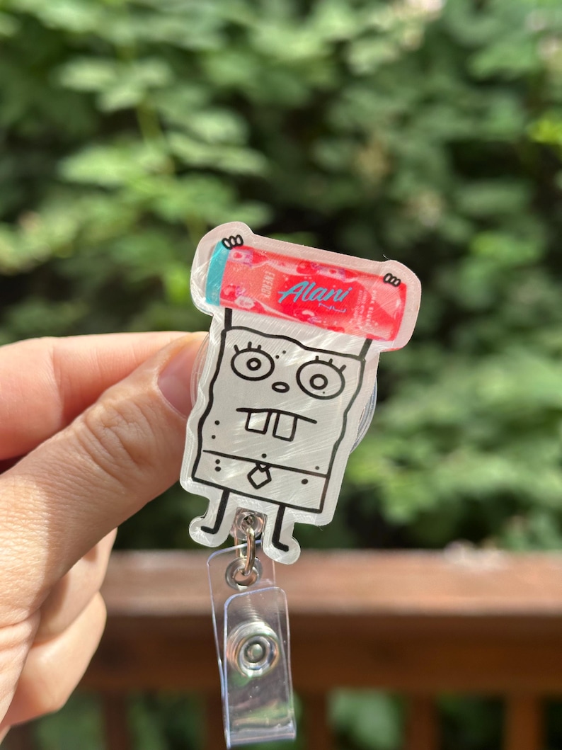 May include: A clear acrylic badge reel featuring a cartoon character design. The character has a square body, large eyes, and a tie. The badge reel has a red and white design with the text "Alani".