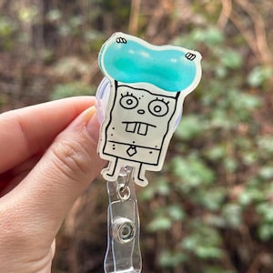 May include: A clear plastic badge holder with a cartoon character design. The character is a light blue, white, and black cartoon with a toothy grin and large eyes. The character is wearing a light blue hat. The badge holder has a retractable cord.
