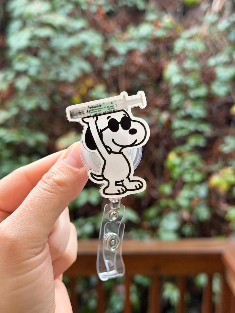May include: A white and black Snoopy badge reel with a syringe design. Snoopy is wearing sunglasses and holding a syringe. The badge reel has a clear plastic clip and a retractable cord.