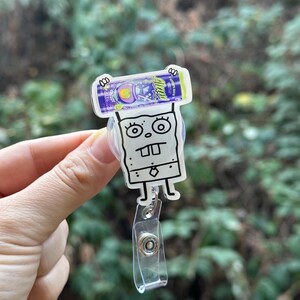 May include: A clear plastic badge reel featuring a cartoon character design. The design includes a purple and blue candy bar and a white cartoon character with a square body and large eyes. The reel has a clear plastic strap and a metal clip.