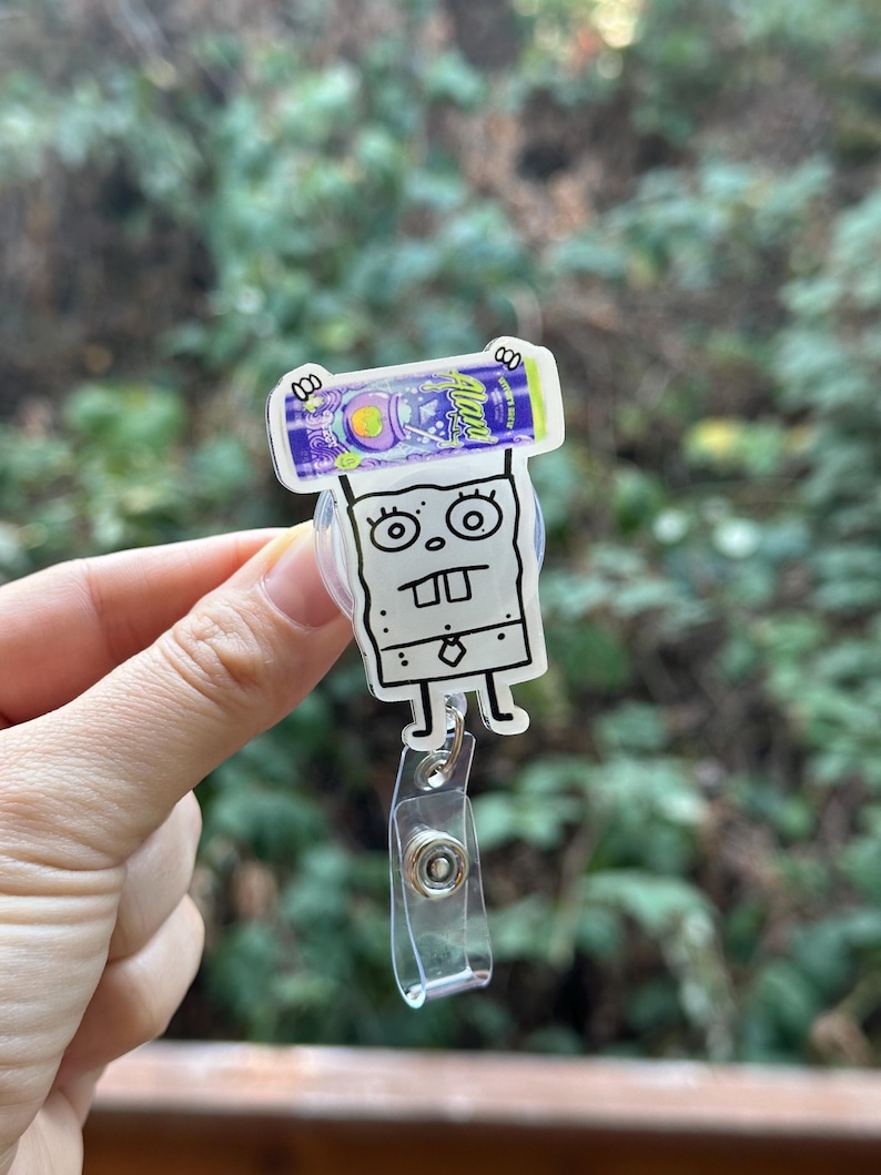 May include: A clear plastic badge reel featuring a cartoon character design. The design includes a purple and blue candy bar and a white cartoon character with a square body and large eyes. The reel has a clear plastic strap and a metal clip.