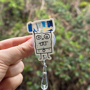 May include: A white cartoon character with a tie, holding a blue and white Ensure Original Nutrition Shake can above its head. The character is attached to a clear plastic retractable badge holder.