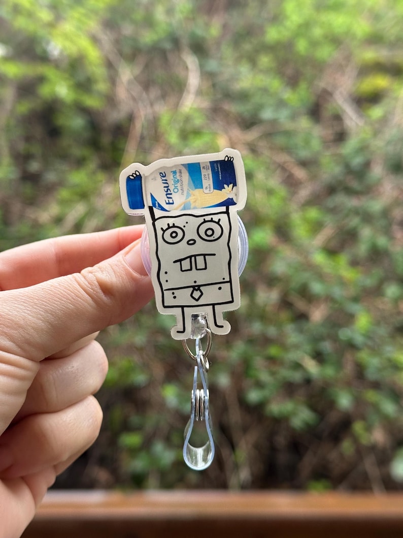 May include: A white cartoon character with a tie, holding a blue and white Ensure Original Nutrition Shake can above its head. The character is attached to a clear plastic retractable badge holder.