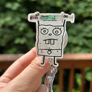 Op de afbeelding: Een heldere acryl badge reel met een stripfiguur met een spuit. De spuit heeft de tekst "Dilaudid" en medische informatie. De reel heeft een doorzichtige plastic clip en een intrekbaar koord.