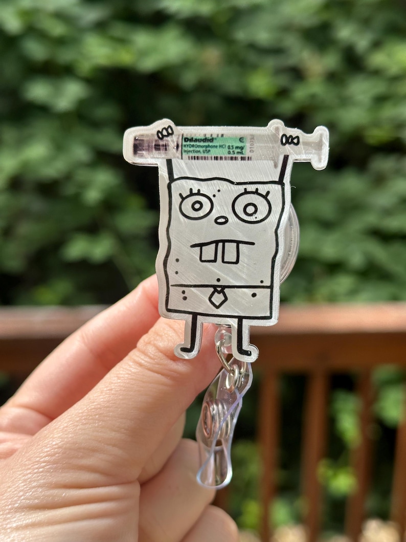 Op de afbeelding: Een heldere acryl badge reel met een stripfiguur met een spuit. De spuit heeft de tekst "Dilaudid" en medische informatie. De reel heeft een doorzichtige plastic clip en een intrekbaar koord.
