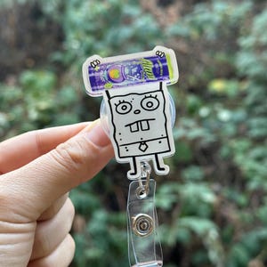 May include: A white and black cartoon badge reel featuring a character with a surprised expression, holding a purple and green beverage can. The reel has a clear plastic strap and a metal clip.