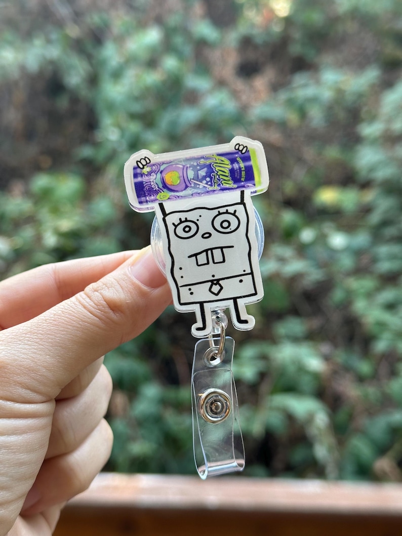May include: A white and black cartoon badge reel featuring a character with a surprised expression, holding a purple and green beverage can. The reel has a clear plastic strap and a metal clip.
