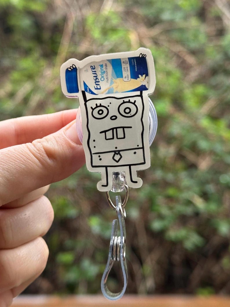 May include: A white cartoon character with a black outline holding a bottle of Ensure Original Nutrition Shake. The character is wearing a tie and has a surprised expression. The character is attached to a clear plastic retractable badge holder.