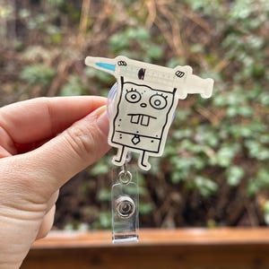 May include: A white cartoon character with a syringe on its head. The character is attached to a clear plastic retractable badge holder.