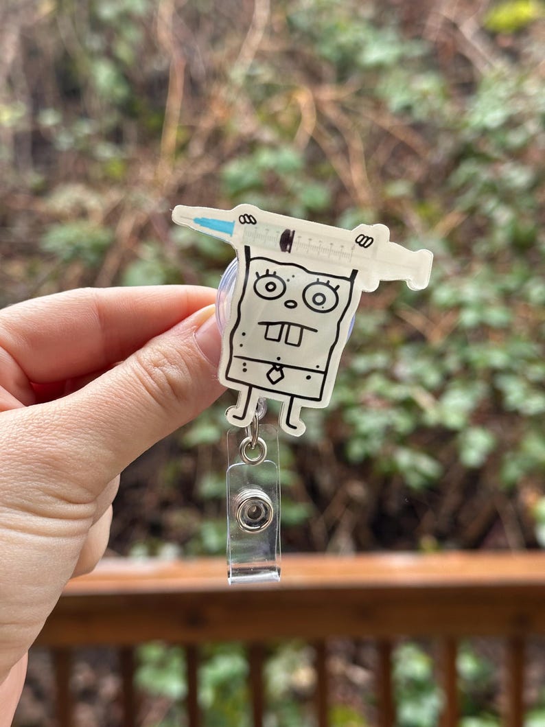 May include: A white cartoon character with a syringe on its head. The character is attached to a clear plastic retractable badge holder.