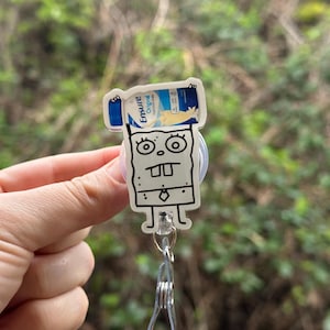 May include: A white cartoon character with a black outline holding a blue and white Ensure Original Nutrition Shake bottle. The character is wearing a tie and has a surprised expression. The character is attached to a clear plastic retractable badge holder.