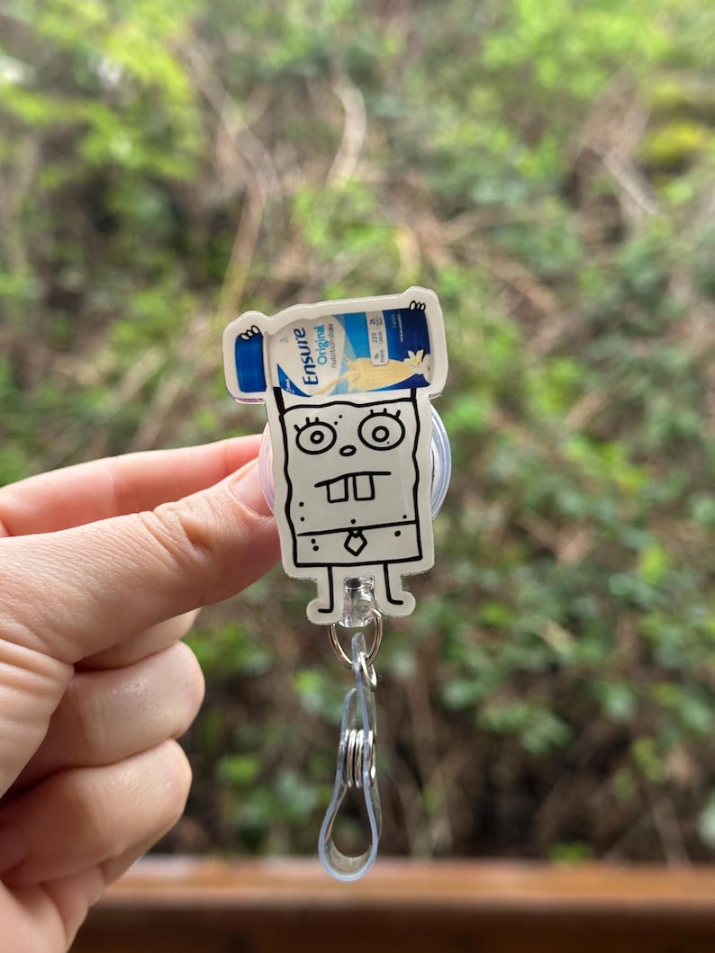 May include: A white cartoon character with a black outline holding a blue and white Ensure Original Nutrition Shake bottle. The character is wearing a tie and has a surprised expression. The character is attached to a clear plastic retractable badge holder.