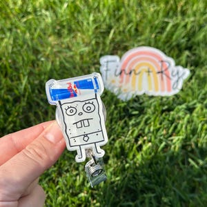 Peut inclure: Un porte-badge en acrylique transparent repr&eacute;sentant un personnage de dessin anim&eacute; avec une canette de Red Bull sur la t&ecirc;te. Le porte-badge a un clip argent&eacute; et un cordon r&eacute;tractable. Un autocollant avec un arc-en-ciel et le texte "Finn + Ry" est en arri&egrave;re-plan.