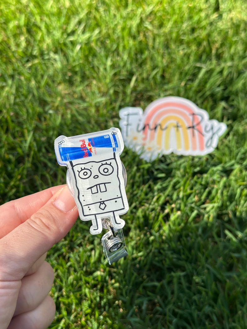 Peut inclure: Un porte-badge en acrylique transparent repr&eacute;sentant un personnage de dessin anim&eacute; avec une canette de Red Bull sur la t&ecirc;te. Le porte-badge a un clip argent&eacute; et un cordon r&eacute;tractable. Un autocollant avec un arc-en-ciel et le texte "Finn + Ry" est en arri&egrave;re-plan.