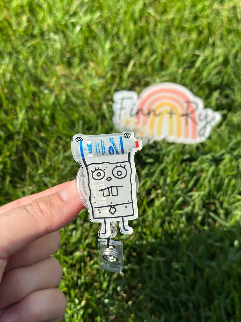 Puede incluir: Un portagafete de pl&aacute;stico transparente con un personaje de dibujos animados que sostiene una botella de desinfectante para manos. El personaje est&aacute; dibujado en blanco y negro y tiene una gran sonrisa. El portagafete tiene un cord&oacute;n retr&aacute;ctil.