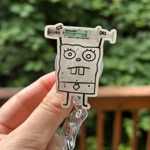 Op de afbeelding: Een doorzichtige badge reel met een stripfiguur en een spuitontwerp. De spuit heeft de tekst "Dilaudid" en andere medische informatie. De reel heeft een doorzichtige plastic clip.
