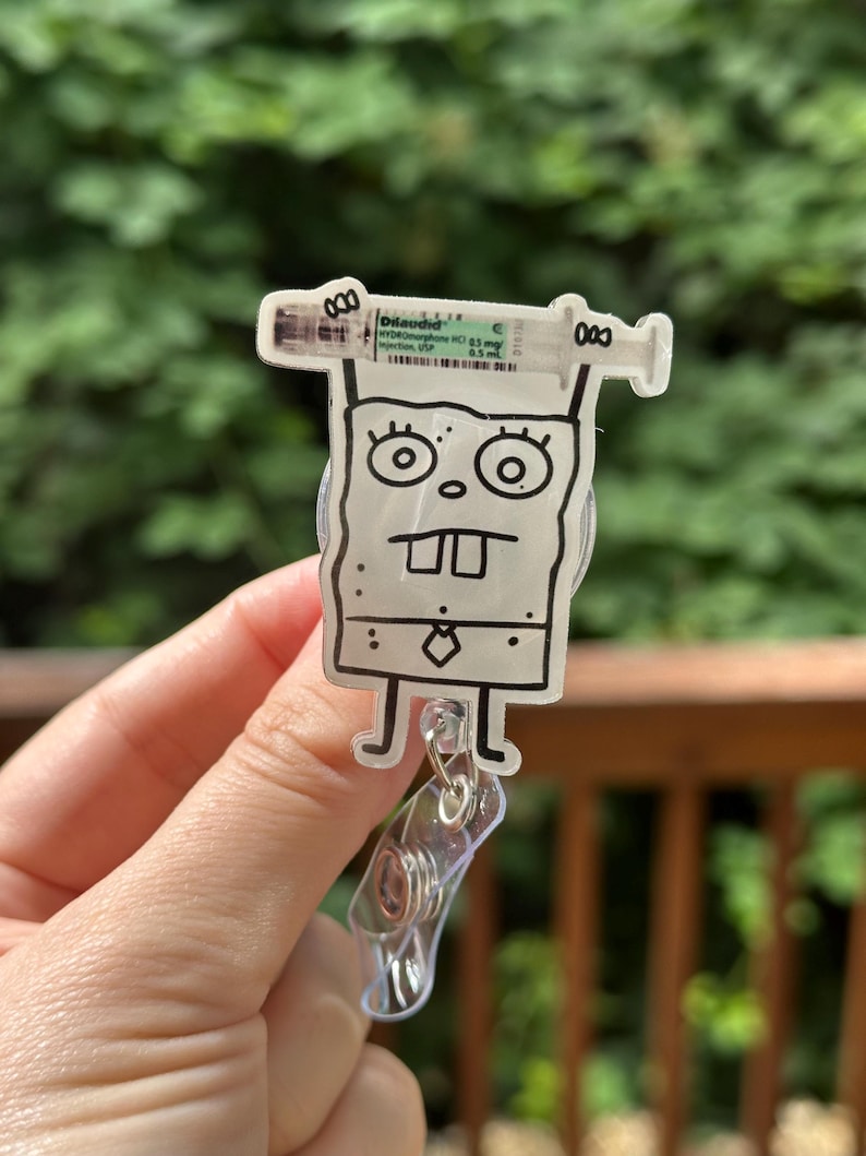 Op de afbeelding: Een doorzichtige badge reel met een stripfiguur en een spuitontwerp. De spuit heeft de tekst "Dilaudid" en andere medische informatie. De reel heeft een doorzichtige plastic clip.