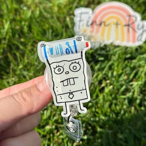 May include: A clear acrylic badge reel featuring a cartoon character holding a bottle of Aquasol 100. The character is a yellow sponge with a black outline and a black tie. The bottle is blue and white with the text "Aquasol 100" printed on it.