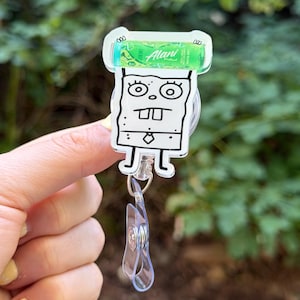 K&ouml;nnte beinhalten: Ein durchsichtiges Acryl-Ausweis-Jojo mit einem Cartoon-Charakter-Design. Die Figur ist eine schwarz-wei&szlig;e Zeichnung mit einem gr&uuml;nen Alani Nu-Produkt oben. Das Jojo hat einen transparenten Kunststoffclip und ein einziehbares Kabel.