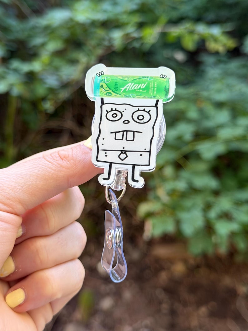 K&ouml;nnte beinhalten: Ein durchsichtiges Acryl-Ausweis-Jojo mit einem Cartoon-Charakter-Design. Die Figur ist eine schwarz-wei&szlig;e Zeichnung mit einem gr&uuml;nen Alani Nu-Produkt oben. Das Jojo hat einen transparenten Kunststoffclip und ein einziehbares Kabel.