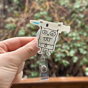 May include: A white cartoon character with a syringe on its head, holding a badge reel. The character has a black outline and is wearing a tie. The badge reel is clear plastic with a silver clip.