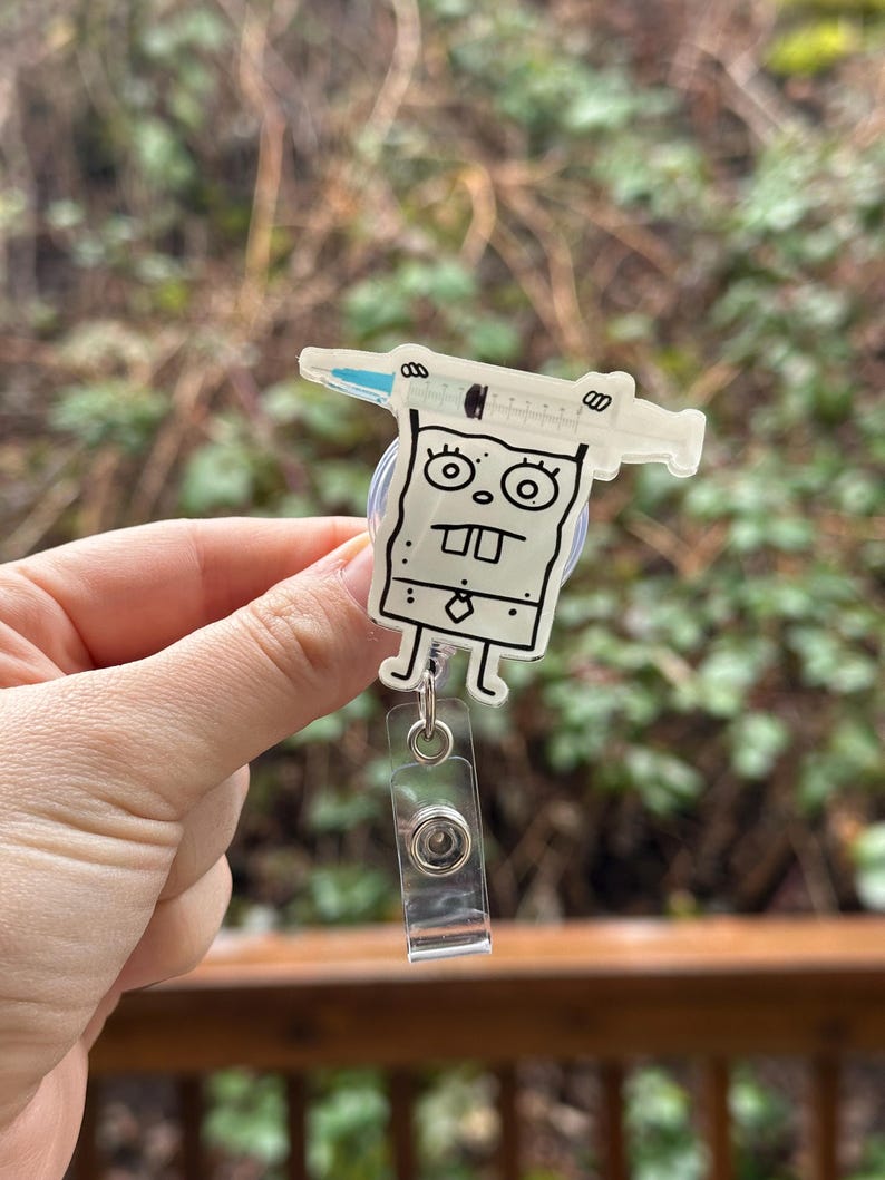 May include: A white cartoon character with a syringe on its head, holding a badge reel. The character has a black outline and is wearing a tie. The badge reel is clear plastic with a silver clip.