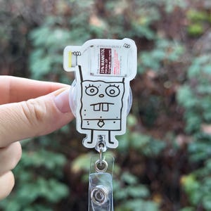 May include: A clear acrylic badge reel featuring a cartoon character design. The design includes a cartoon character with a bottle of 25% Mannitol injection. The badge reel has a clear plastic strap and a metal clip.