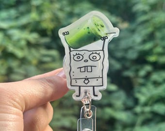 May include: A clear acrylic badge reel featuring a cartoon character holding a green beverage. The character is outlined in black with a white body and a clear plastic clip and reel attached.