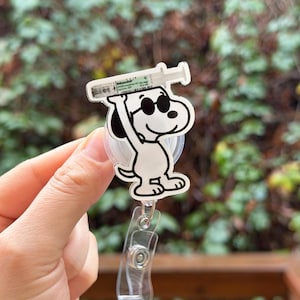 May include: A white and black Snoopy badge reel with a syringe graphic. The cartoon Snoopy is wearing sunglasses and holding the syringe above his head. The badge reel has a clear plastic clip and a retractable cord.