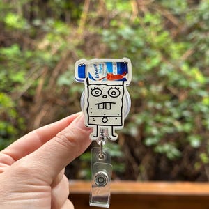 May include: A clear plastic badge holder with a cartoon character holding a bottle of Ensure Original nutritional drink. The character is white with black outlines and has a surprised expression. The badge holder has a retractable cord.