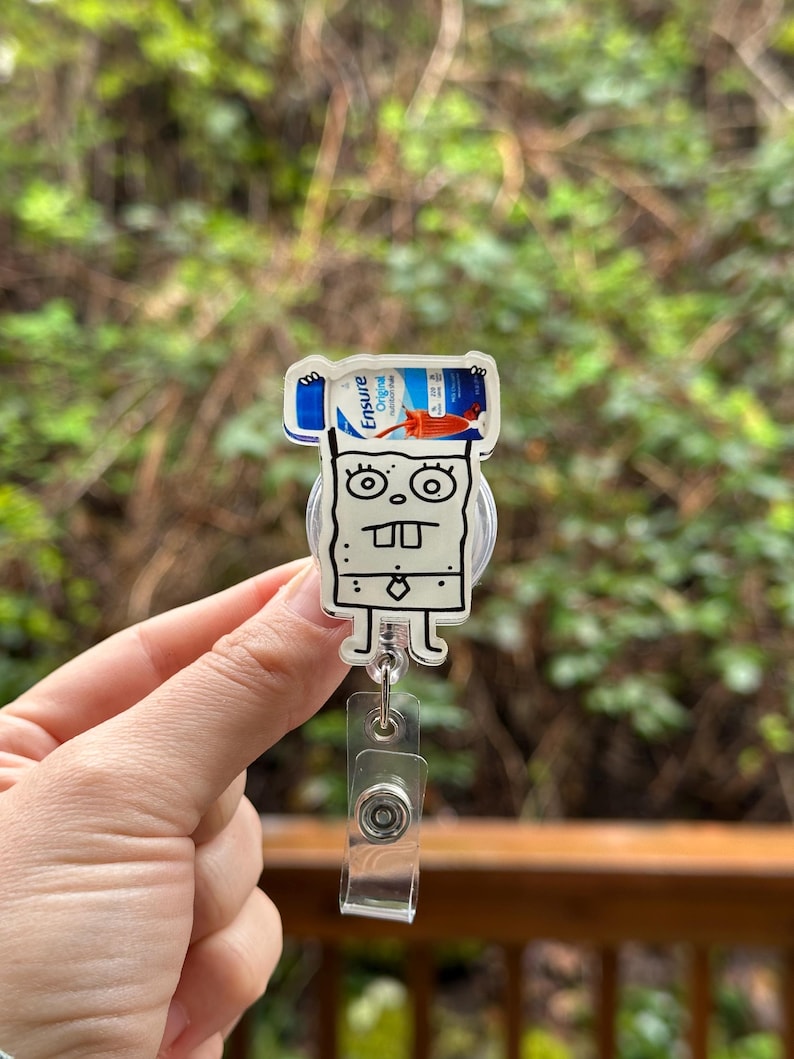 May include: A clear plastic badge holder with a cartoon character holding a bottle of Ensure Original nutritional drink. The character is white with black outlines and has a surprised expression. The badge holder has a retractable cord.