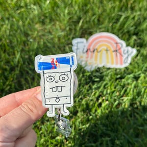 Peut inclure: Un porte-badge en acrylique transparent repr&eacute;sentant un personnage de dessin anim&eacute; avec deux canettes de boisson &eacute;nergisante Red Bull sur le dessus. Le porte-badge a un clip en plastique transparent et un cordon r&eacute;tractable. L'arri&egrave;re-plan est de l'herbe verte floue.