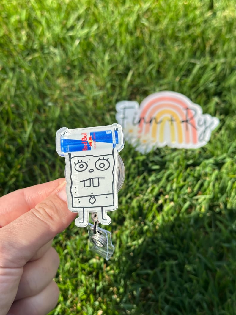 Peut inclure: Un porte-badge en acrylique transparent repr&eacute;sentant un personnage de dessin anim&eacute; avec deux canettes de boisson &eacute;nergisante Red Bull sur le dessus. Le porte-badge a un clip en plastique transparent et un cordon r&eacute;tractable. L'arri&egrave;re-plan est de l'herbe verte floue.