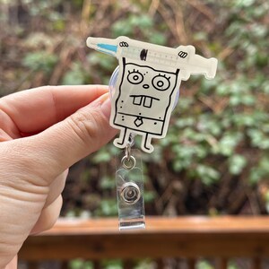 May include: A clear plastic badge holder with a cartoon character design. The character is a square with a face and a syringe on top of its head. The badge holder has a retractable clip.