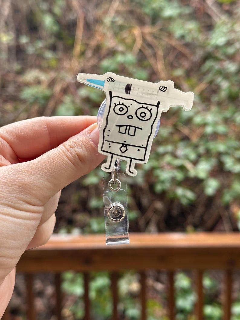 May include: A clear plastic badge holder with a cartoon character design. The character is a square with a face and a syringe on top of its head. The badge holder has a retractable clip.