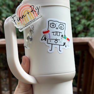 May include: A white tumbler with a handle, featuring two custom badge reels. One reel has a rainbow design with the text "Finn + Ry" and "CUSTOM BADGE REELS MADE TO BRIGHTEN UP YOUR WORKPLACE!". The other reel has a cartoon character.