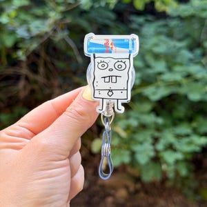May include: A clear acrylic badge reel featuring a cartoon character with a square face, large eyes, and buck teeth. The reel holds two small Red Bull energy drink cans. A silver-colored clip and a clear plastic loop are attached.