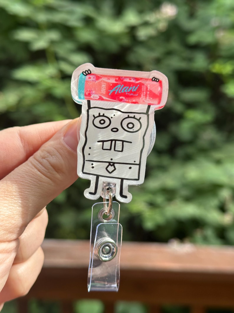 May include: A clear acrylic badge reel featuring a cartoon character with a red and blue energy drink. The badge reel has a clear plastic clip and a retractable cord. The energy drink has the word "Alani" on it.