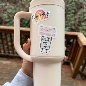 May include: A cream-colored travel mug with a handle, featuring two stickers. One sticker has a rainbow and the text "Finn + Ry." The other sticker depicts a cartoon character holding an EKG graph.