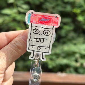 May include: A clear acrylic badge reel featuring a cartoon character with a red and white energy drink. The character has a white body, black outline, and a tie. The reel has a clear plastic clip.