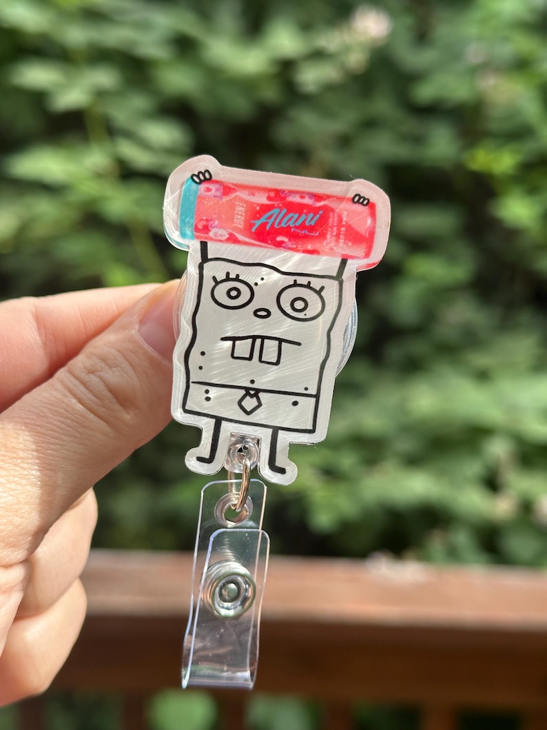 May include: A clear acrylic badge reel featuring a cartoon character with a red and white energy drink. The character has a white body, black outline, and a tie. The reel has a clear plastic clip.