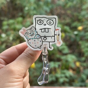 May include: A clear acrylic badge reel featuring a cartoon character holding a baby. The cartoon character is outlined in black with a white background. The baby is wearing a pink bow and a patterned blanket. The reel has a clear plastic clip.