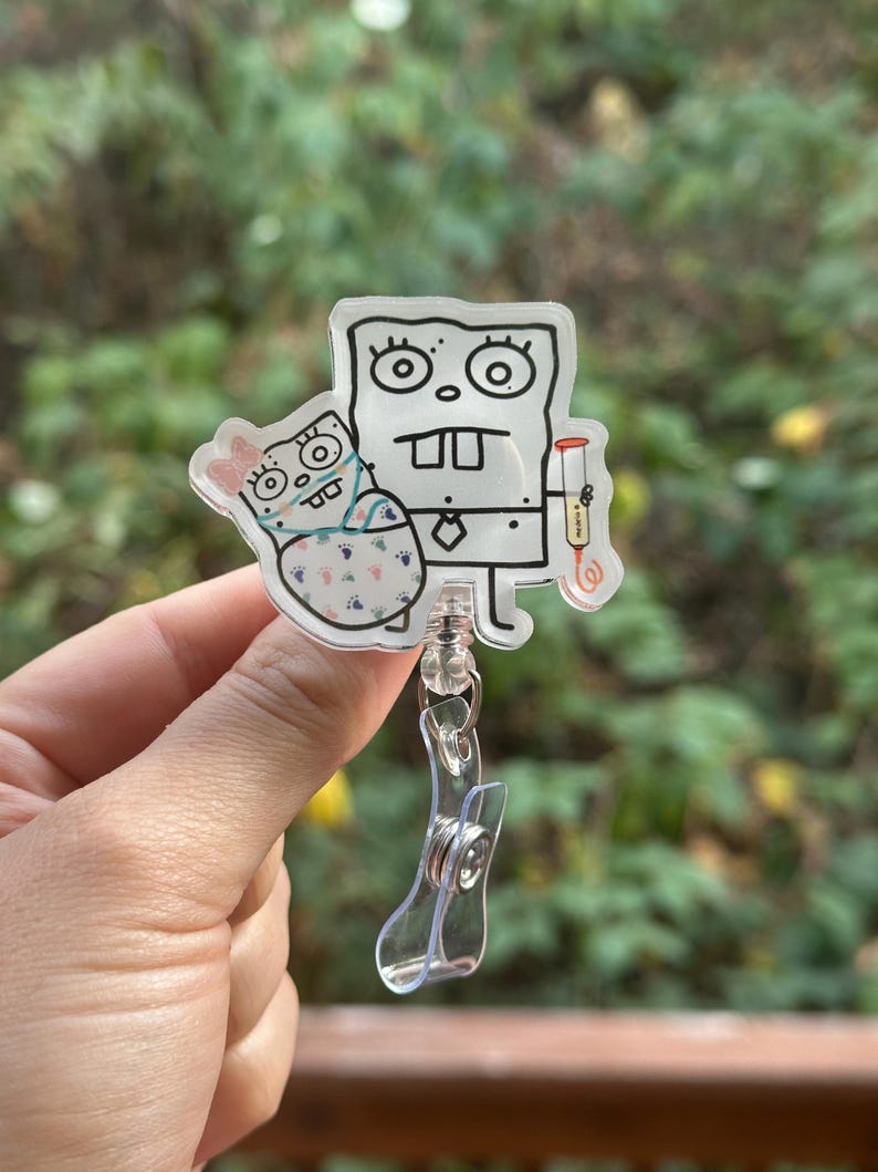 May include: A clear acrylic badge reel featuring a cartoon character holding a baby. The cartoon character is outlined in black with a white background. The baby is wearing a pink bow and a patterned blanket. The reel has a clear plastic clip.
