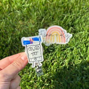 Peut inclure: Un porte-badge en acrylique transparent avec un personnage de dessin anim&eacute; et une canette de Red Bull sur le dessus. Le porte-badge a un cordon r&eacute;tractable et un clip. Un deuxi&egrave;me acrylique transparent avec un arc-en-ciel et le texte "Finn + Ry" est &eacute;galement visible.