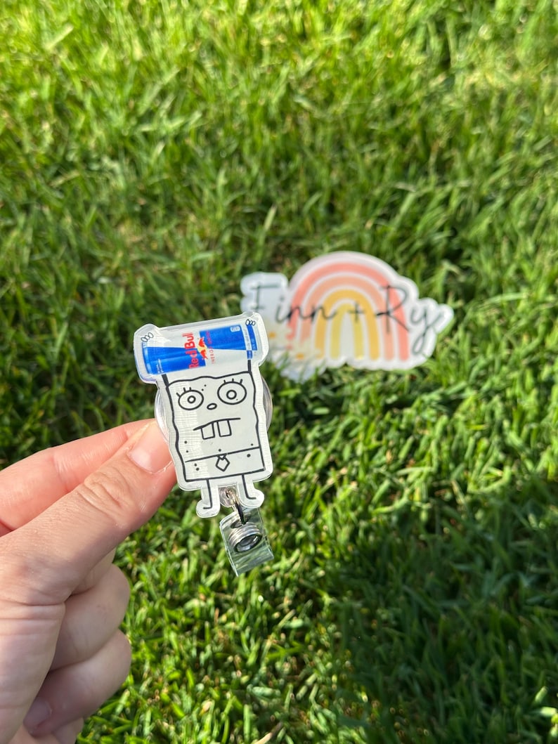Peut inclure: Un porte-badge en acrylique transparent avec un personnage de dessin anim&eacute; et une canette de Red Bull sur le dessus. Le porte-badge a un cordon r&eacute;tractable et un clip. Un deuxi&egrave;me acrylique transparent avec un arc-en-ciel et le texte "Finn + Ry" est &eacute;galement visible.
