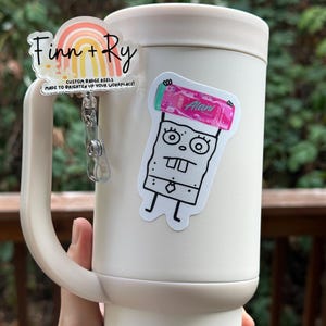 May include: A cream-colored tumbler with a handle, adorned with two custom badge reels. One reel features a rainbow design with the text "Finn + Ry" and "CUSTOM BADGE REELS MADE TO BRIGHTEN UP YOUR WORKPLACE!". The other reel has a cartoon character.