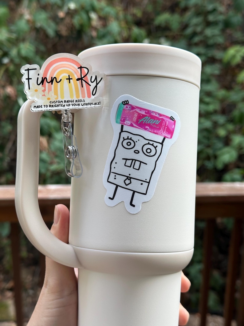 May include: A cream-colored tumbler with a handle, adorned with two custom badge reels. One reel features a rainbow design with the text "Finn + Ry" and "CUSTOM BADGE REELS MADE TO BRIGHTEN UP YOUR WORKPLACE!". The other reel has a cartoon character.