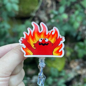 May include: A red and yellow cartoon character with a surprised expression, set on fire, is attached to a clear plastic retractable badge holder.