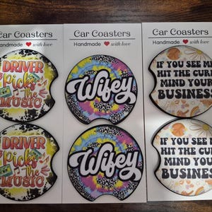 May include: Six circular car coasters arranged in pairs on three white display cards. The coasters feature colorful designs with text including Driver Picks the Music, Wifey, and If You See Me Hit the Curb Mind Your Business, along with graphics.