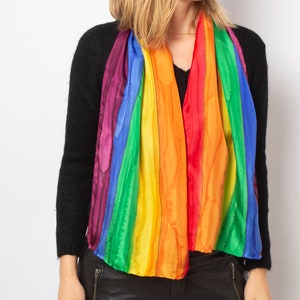 May include: A colorful rainbow scarf with vertical stripes of red, orange, yellow, green, blue, and purple. The scarf is draped over a black sweater. The scarf is made of a lightweight, silky material.